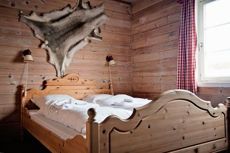 Et rom med trevegger, dobbeltseng i tre og et reinskinn hengende på veggen over sengen.