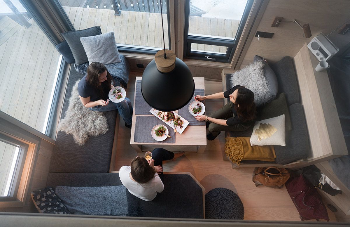 Bilde tatt ovenfra ned som viser tre personer rundt et bord i en liten hytte med mye vinduer rundt