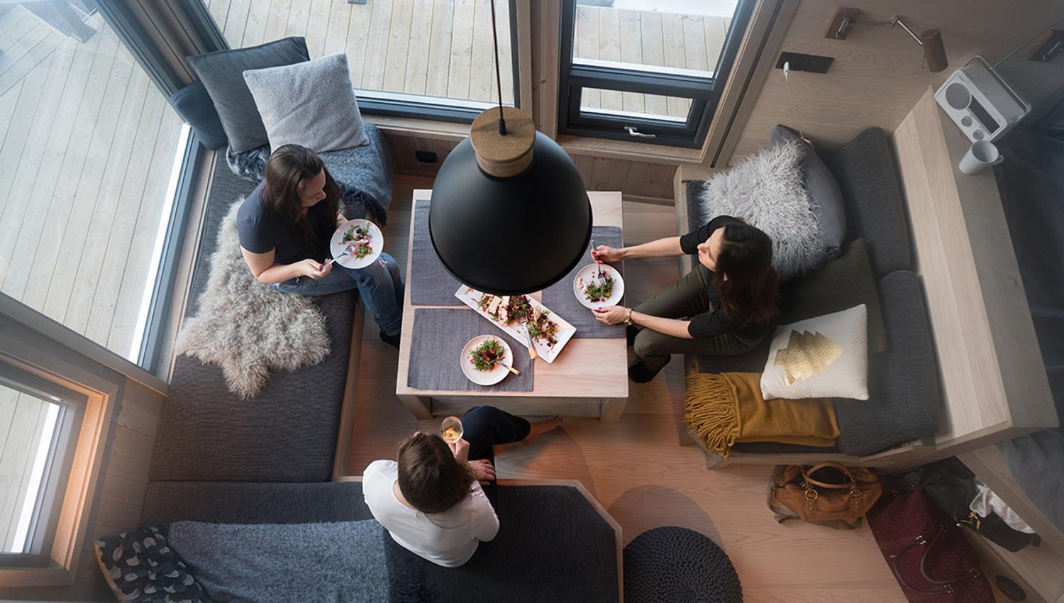 Bilde tatt ovenfra ned som viser tre personer rundt et bord i en liten hytte med mye vinduer rundt