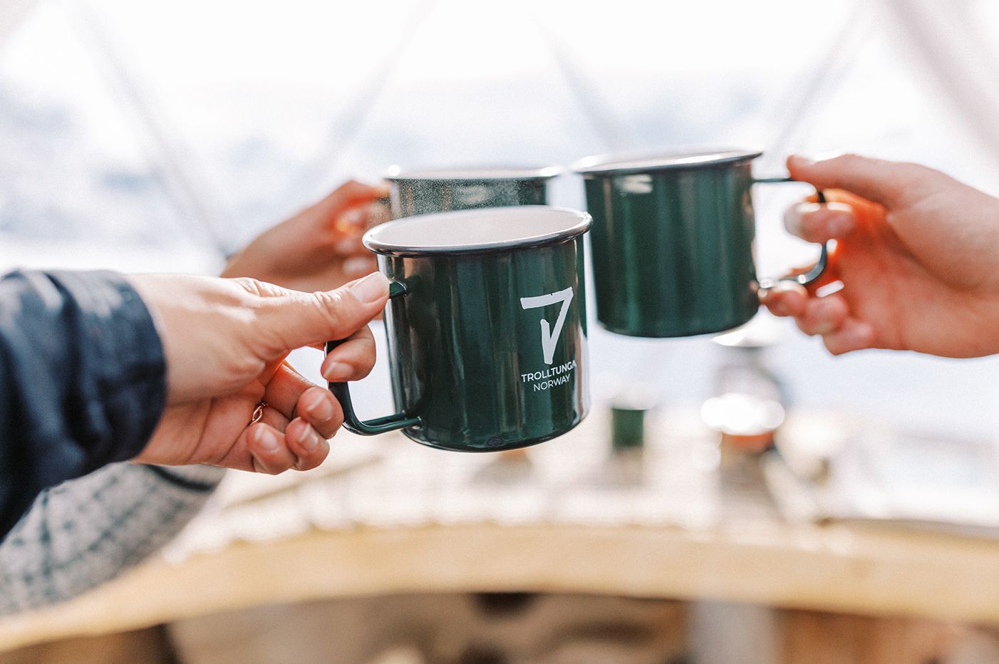 Two individuals clinking green enamel mugs labeled "Trolltunga Norway" in a cozy outdoor setting.