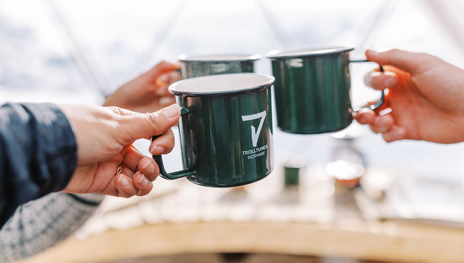 Two individuals clinking green enamel mugs labeled "Trolltunga Norway" in a cozy outdoor setting.