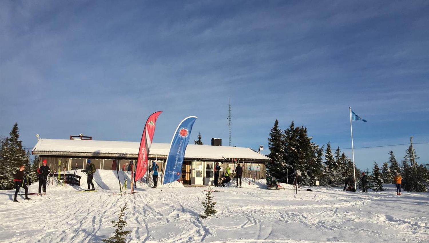 En vinterlig hytte eller fjellstue omgitt av snødekt terreng, med flagg og beachflagg som vaier foran bygningen under klar himmel.