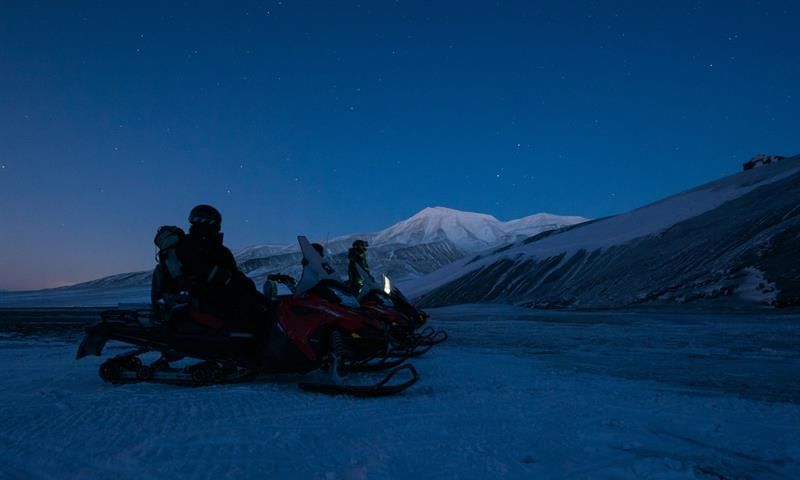 Two snowmobiles with drivers standing on snowcovered landscape during the polar night