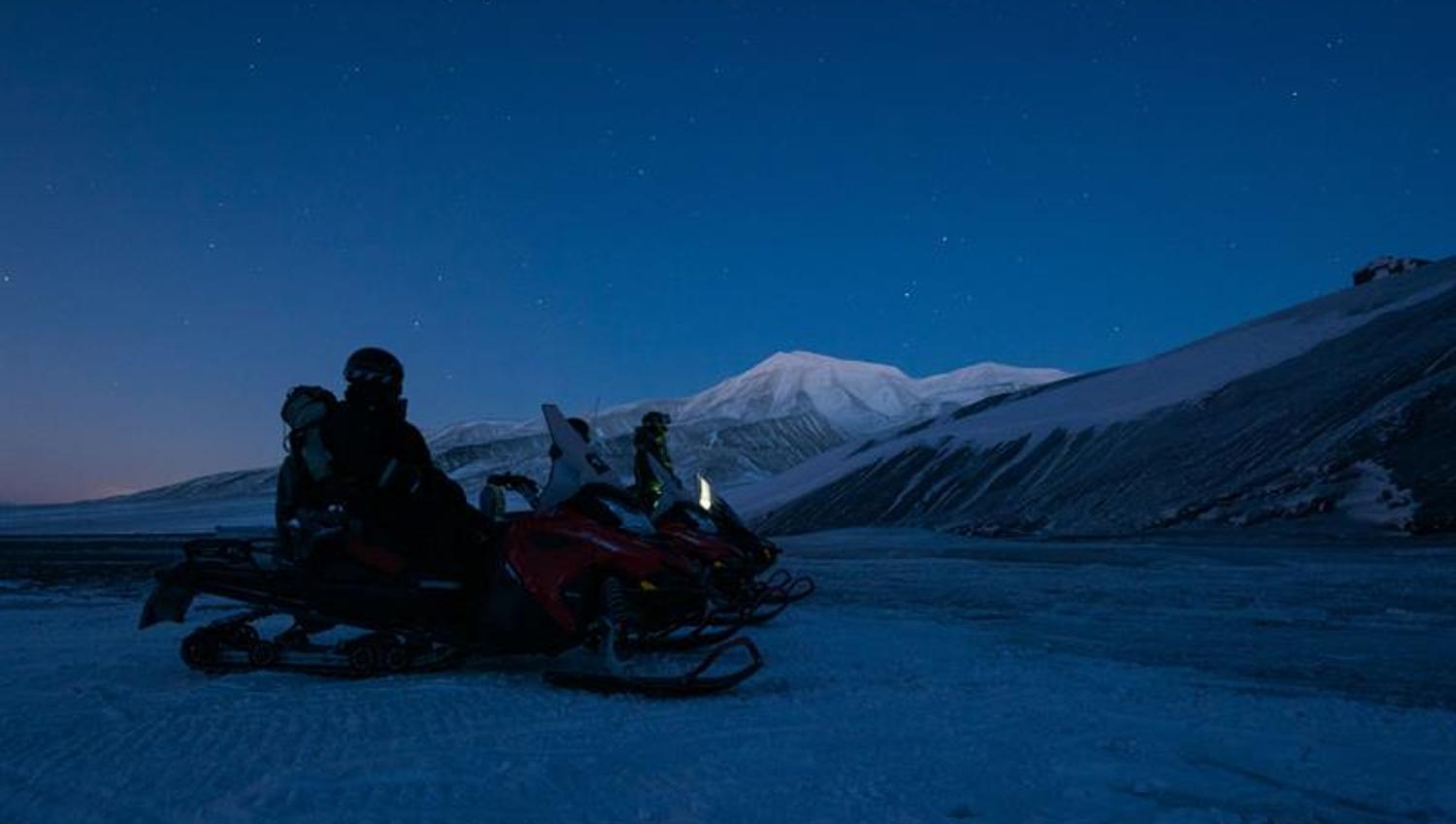 Two snowmobiles with drivers standing on snowcovered landscape during the polar night