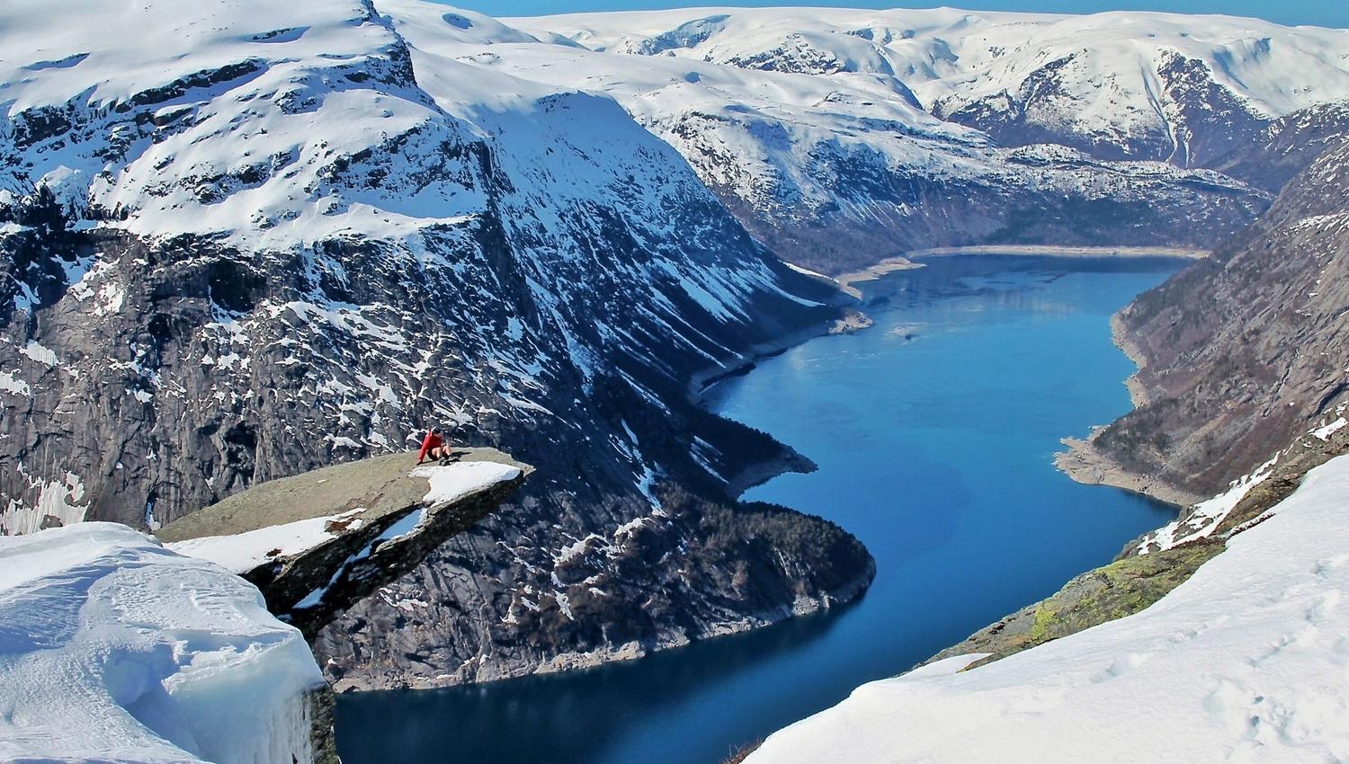 Person sit på Trolltunga om vinteren med utsikt over den snødekte fjorden og fjellandskapet.