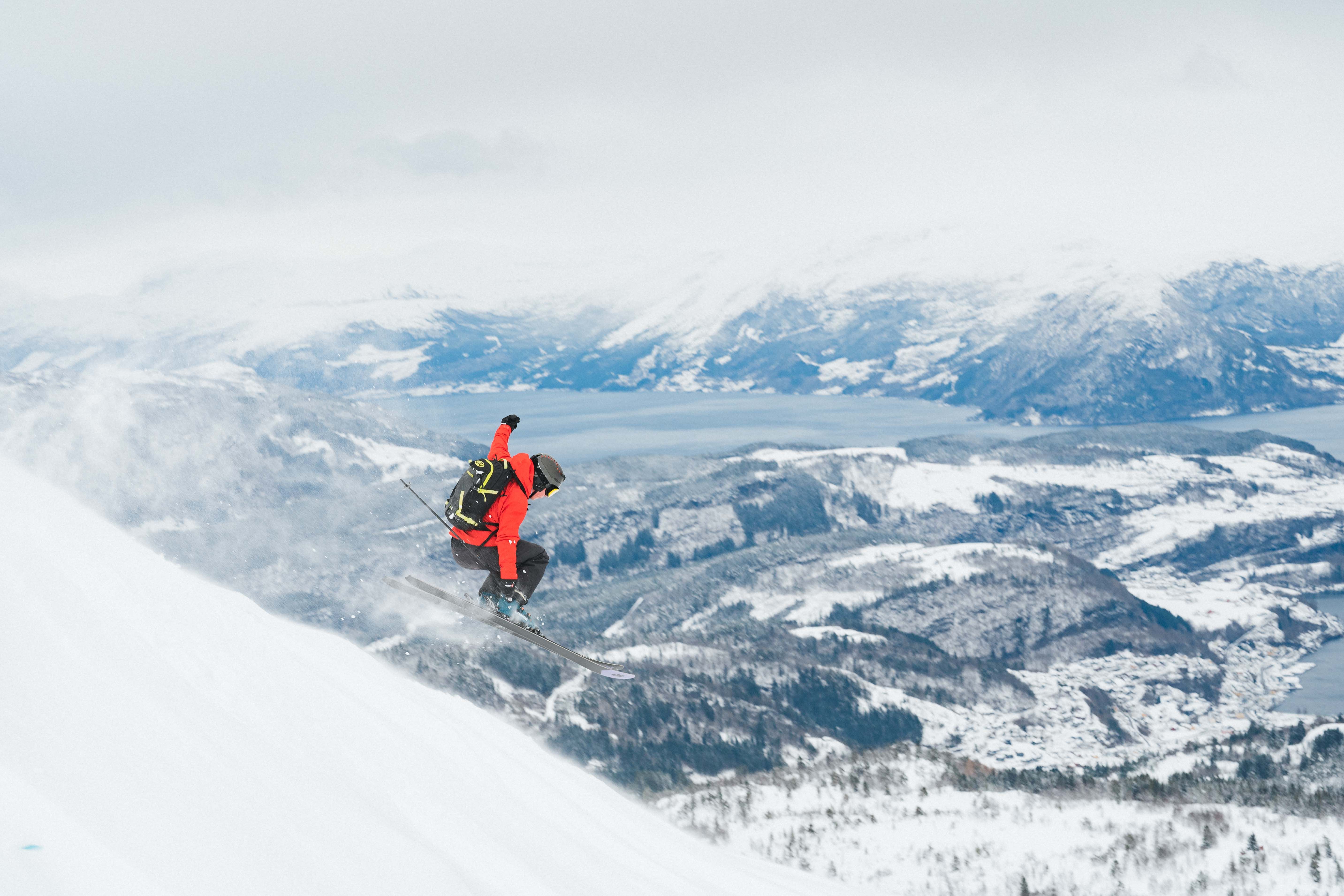 En person med rød jakke hopper utfor en skråning på ski. Utsikt til fjord og snødekket landskap i bakgrunnen.