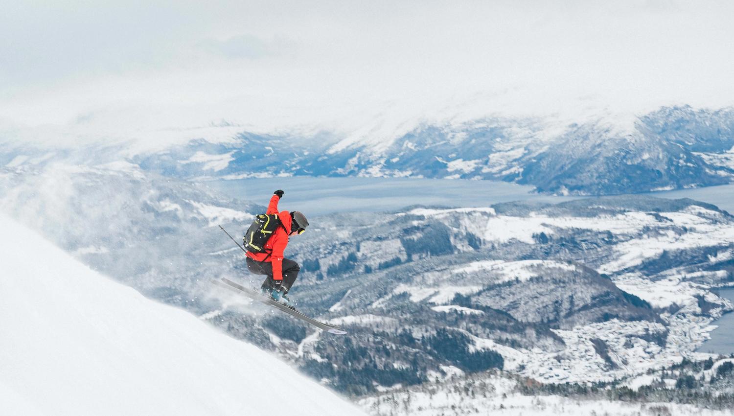 En person med rød jakke hopper utfor en skråning på ski. Utsikt til fjord og snødekket landskap i bakgrunnen.