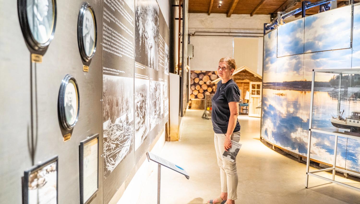 Bildet viser en kvinne som ser på en utstilling på et museum. Hun står foran en vegg med gamle bilder og tekst, og holder en vannflaske i hånden. I bakgrunnen sees en del av en utstilling med bilder av vann og tømmerstokker.