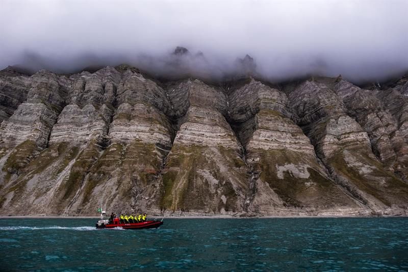 Båt kjører i fjorden med en turgruppe. Bratte fjell i bakgrunnen. Skyer henger over fjellene.