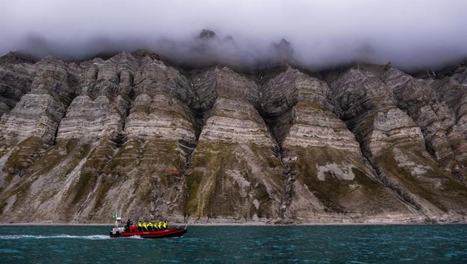 Båt kjører i fjorden med en turgruppe. Bratte fjell i bakgrunnen. Skyer henger over fjellene.