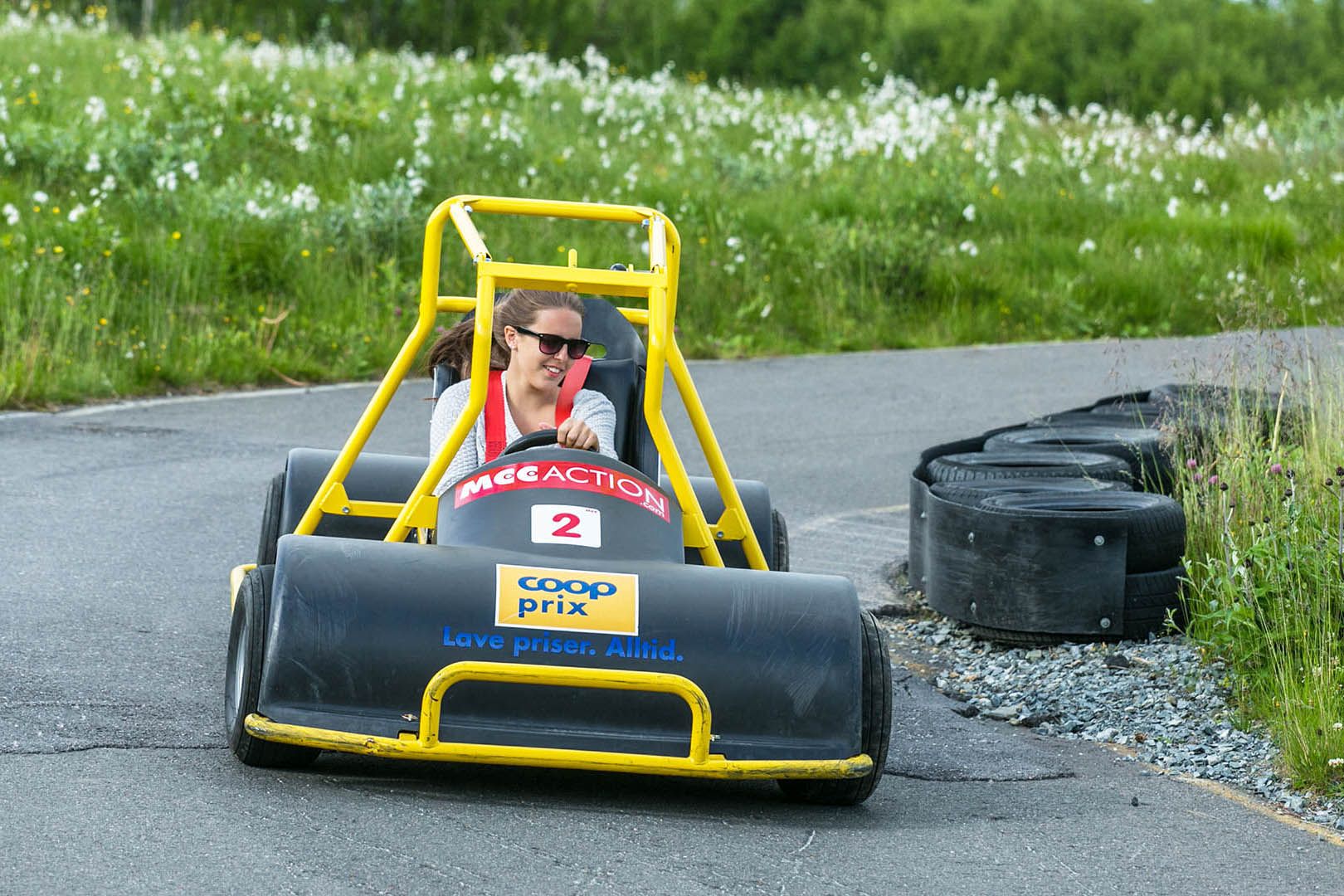 En jente med solbriller kjører en olabil ned en asfaltert downhill-løype gjennom en blomstrende myrulleng.