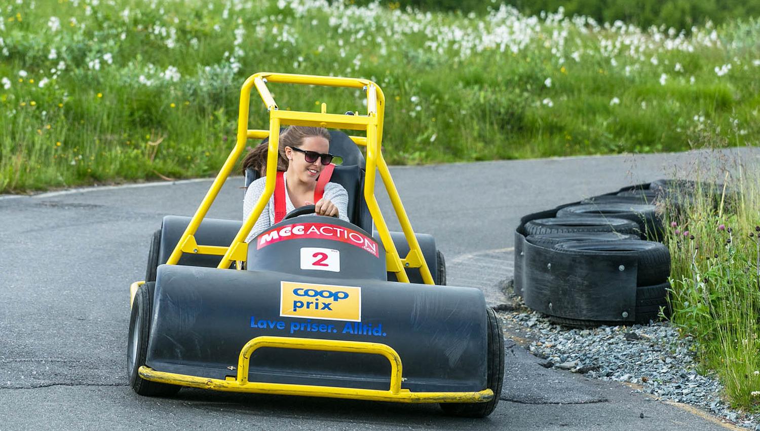 En jente med solbriller kjører en olabil ned en asfaltert downhill-løype gjennom en blomstrende myrulleng.