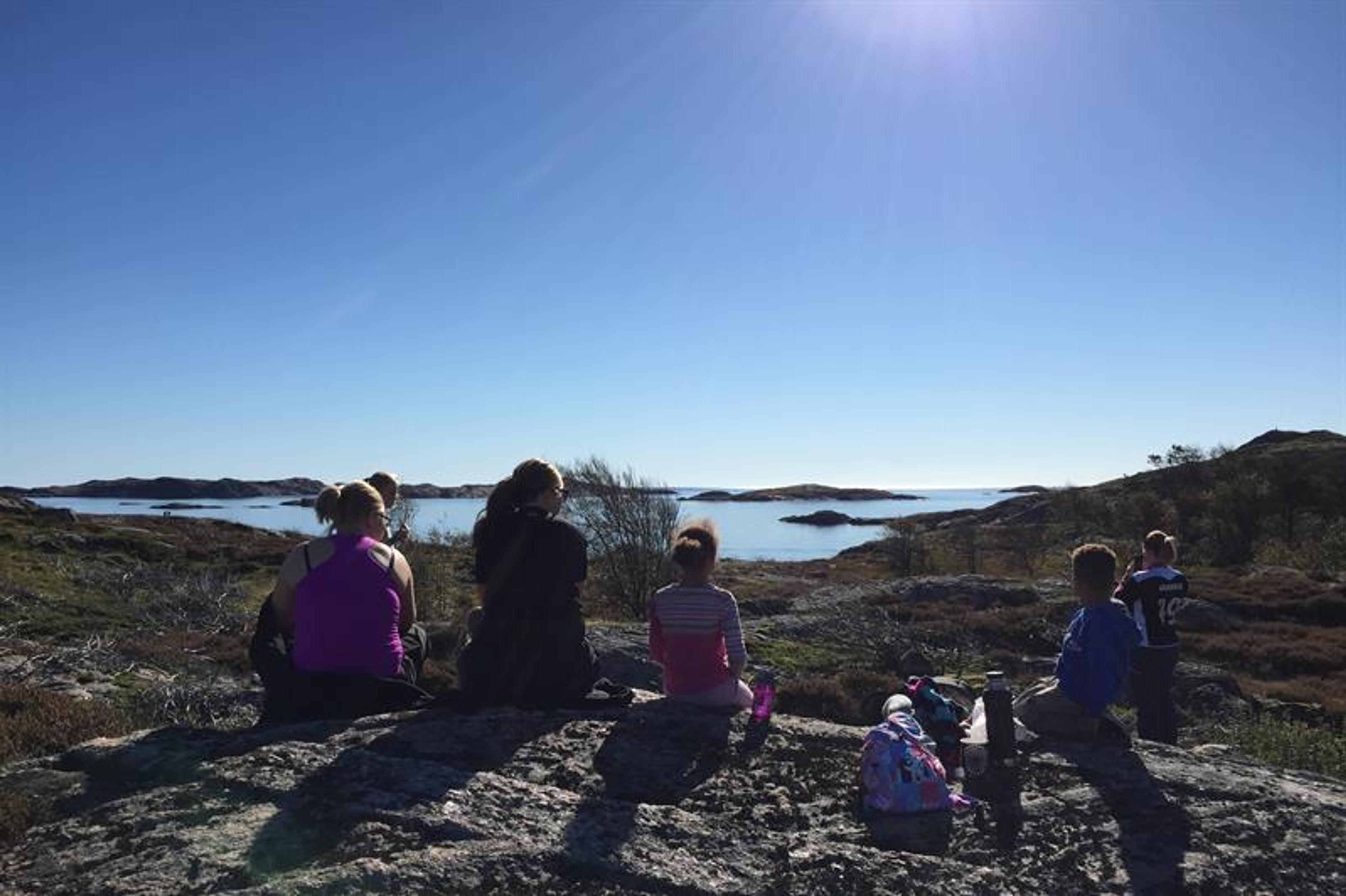 Bilde av seks mennesker som sitter på stein og ser utover mot havet, sola skinner ned på dem, en dame tar bilde av utsikten