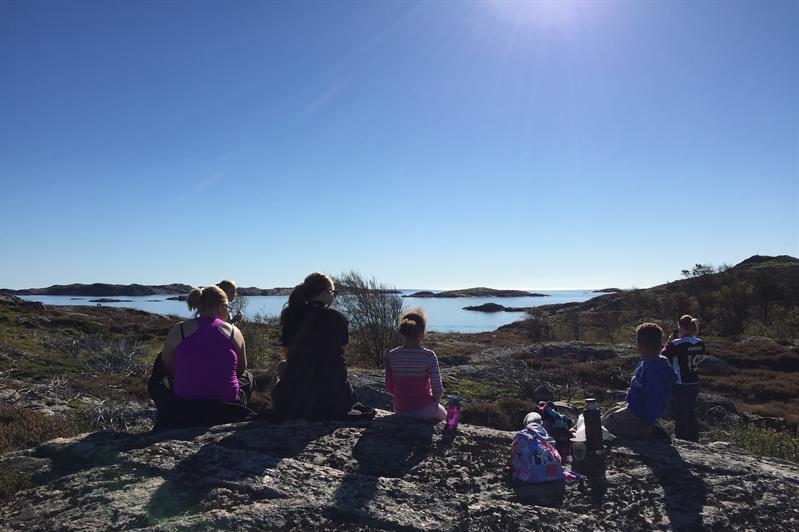 Bilde av seks mennesker som sitter på stein og ser utover mot havet, sola skinner ned på dem, en dame tar bilde av utsikten