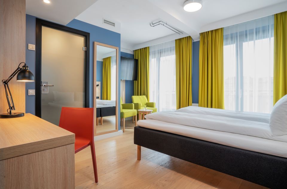 Room interior: Double bed, desk and chair, green armchairs, blue-painted walls and yellow curtains.