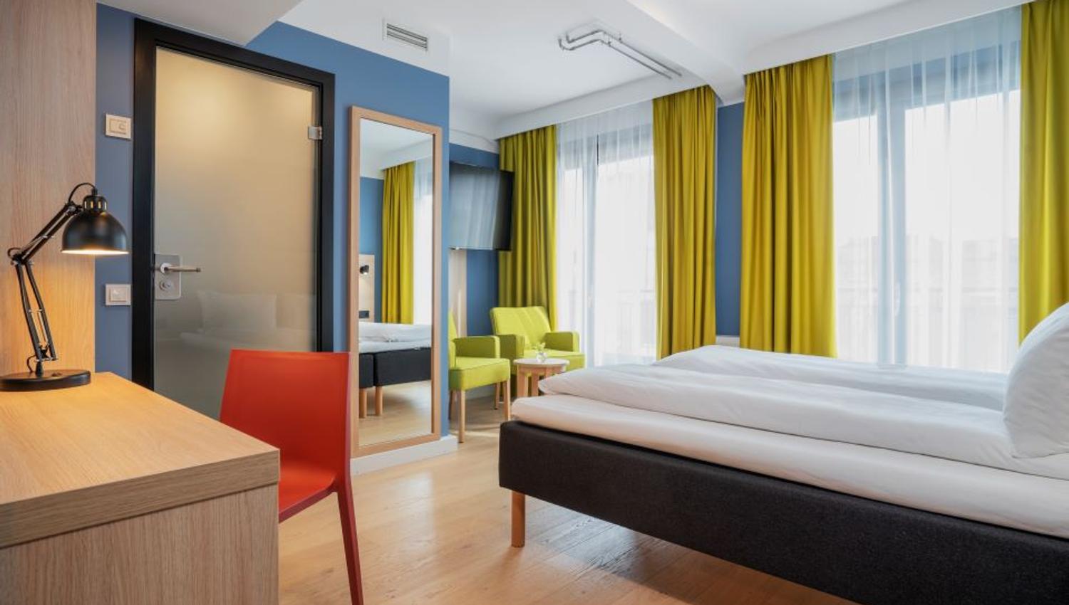 Room interior: Double bed, desk and chair, green armchairs, blue-painted walls and yellow curtains.
