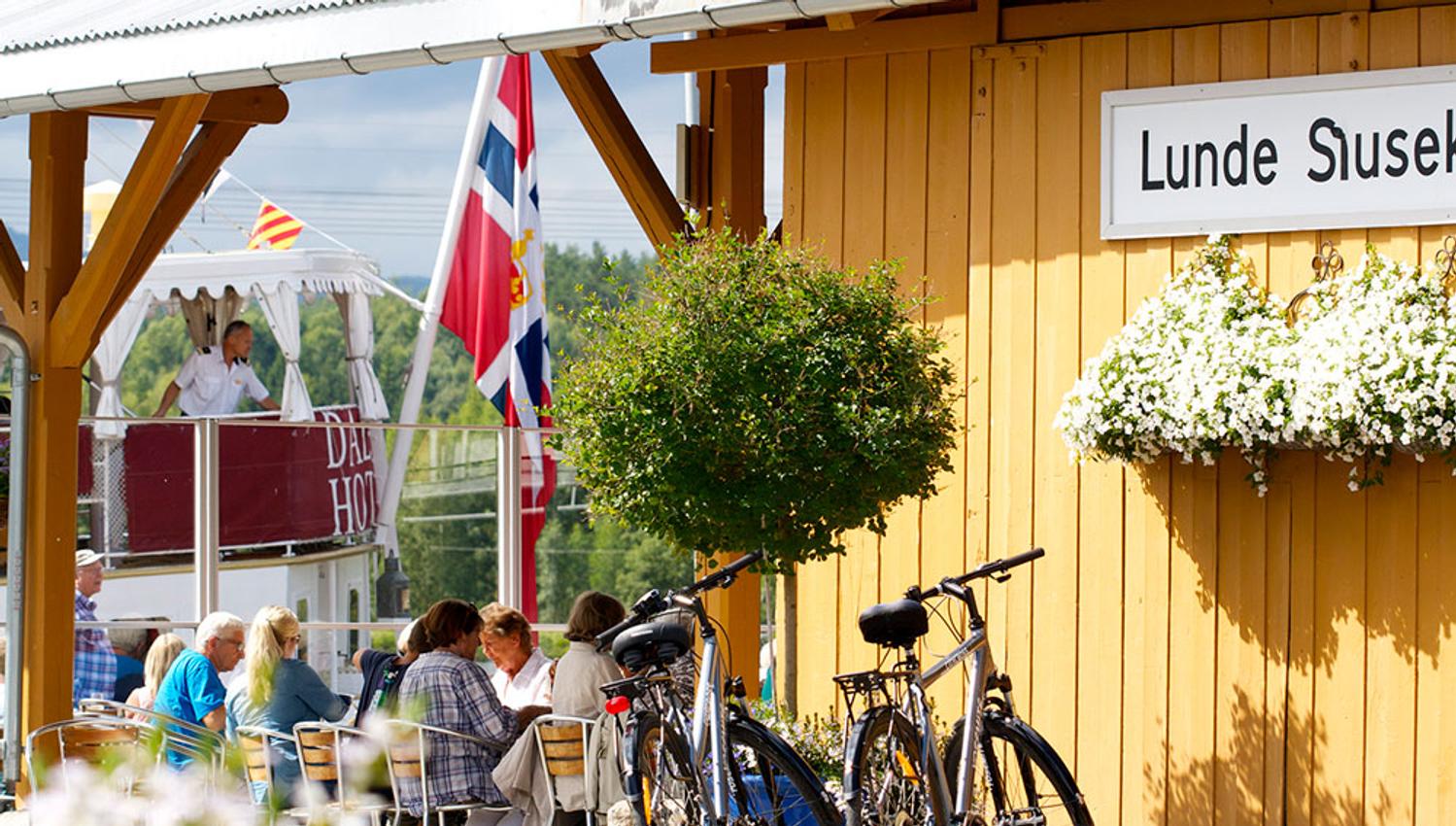 folk som sitter og spiser på Lunde Slusekro mens kanalbåten MS Henrik Ibsen seiler forbi