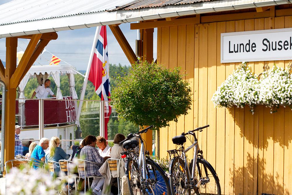 folk som sitter og spiser på Lunde Slusekro mens kanalbåten MS Henrik Ibsen seiler forbi