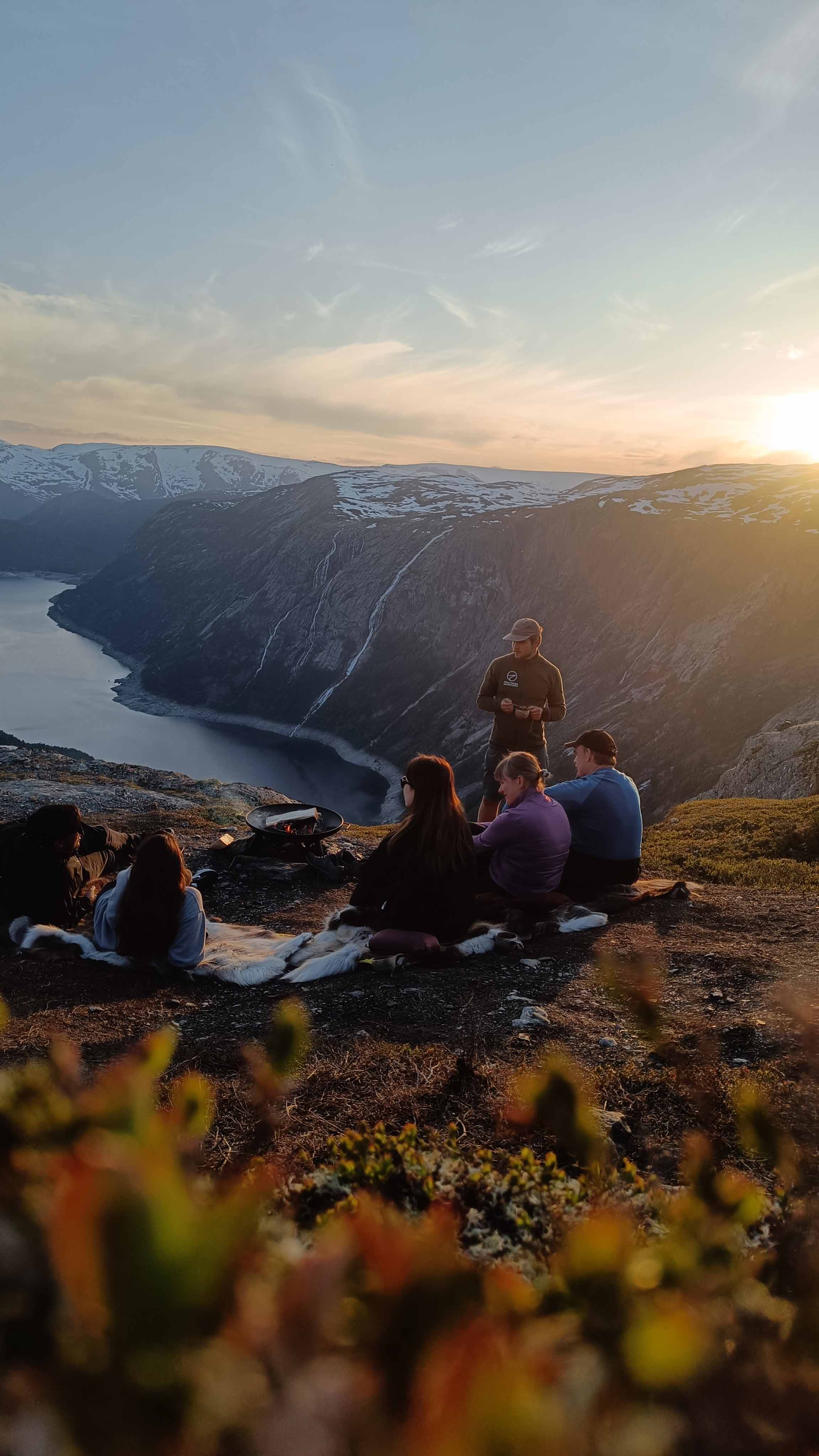 En gruppe mennesker sitter på fjellet og ser på solnedgangen over en fjord.