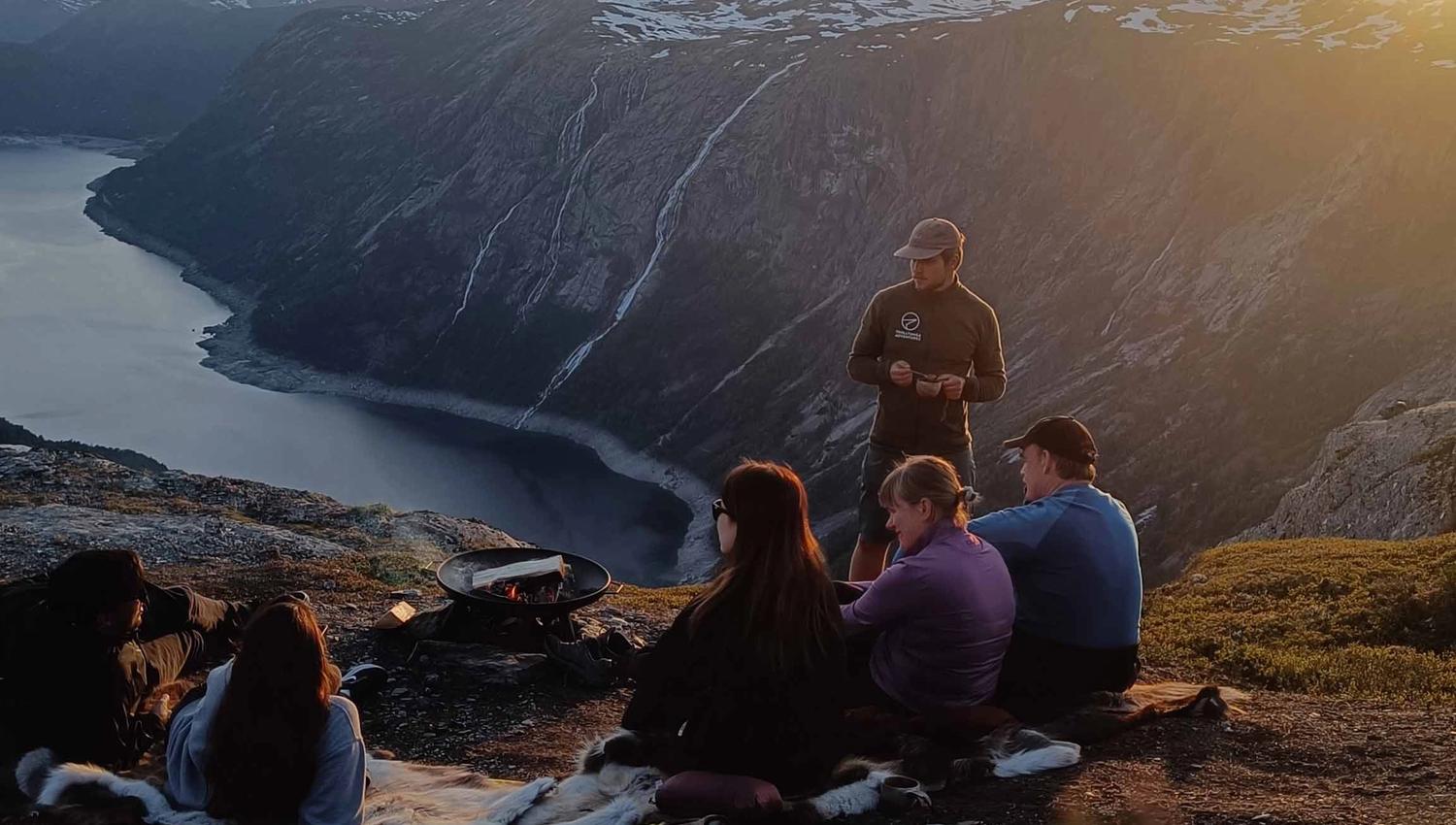 En gruppe mennesker sitter på fjellet og ser på solnedgangen over en fjord.