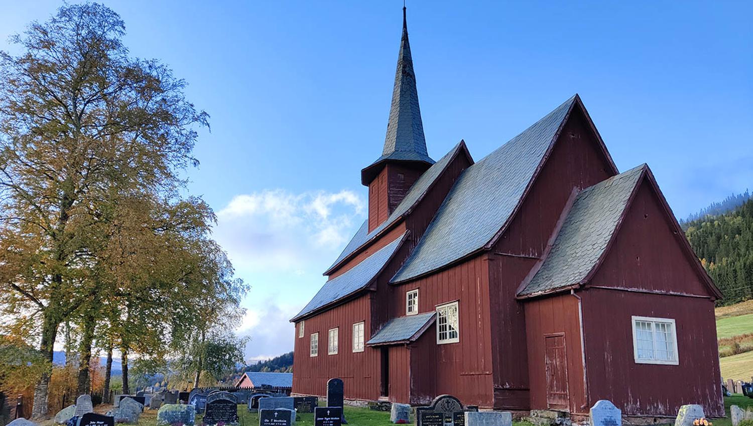 A red-brown stave church surrounded by a church yard with birch trees that start to get autumn-coloured leaves.