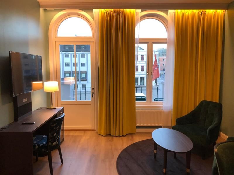 Nyoppusset rom Thon Hotel Høyers med gule gardiner som går fra gulv til tak, 2 buende vinduer, en stol og et ovalt bord, skrivepult og et tv.
