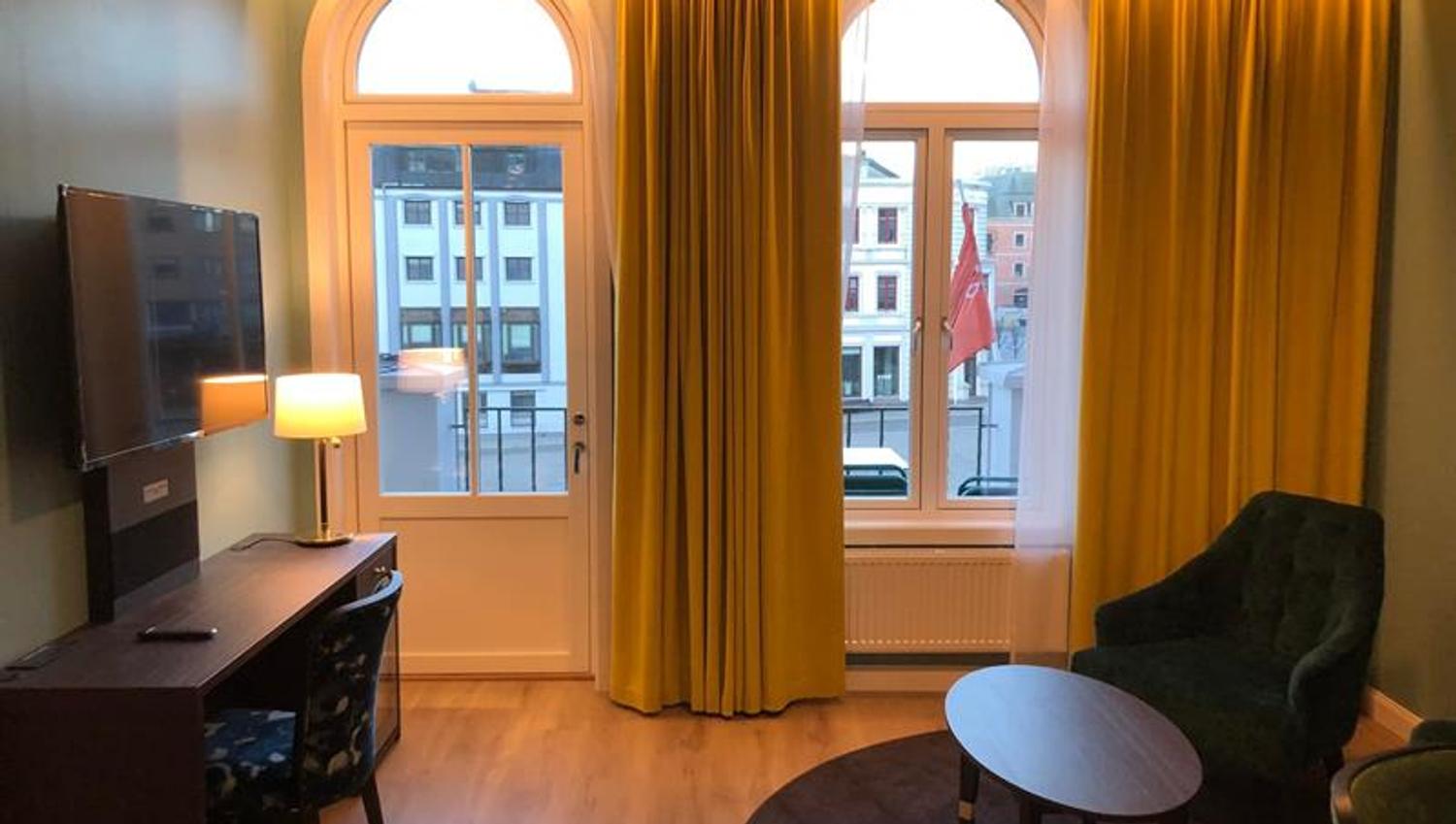 Nyoppusset rom Thon Hotel Høyers med gule gardiner som går fra gulv til tak, 2 buende vinduer, en stol og et ovalt bord, skrivepult og et tv.