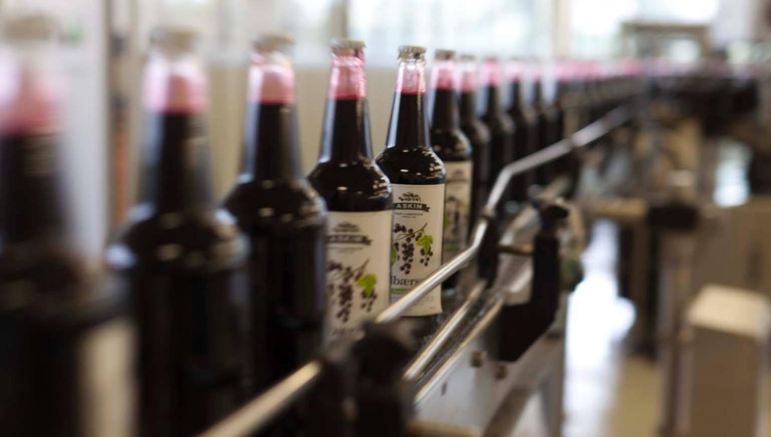 Flasker med mørkerød saft står på rekke og rad på et samlebånd i et produksjonslokale. Etiketten viser druer, og lyset fra vinduene skinner på flaskene.