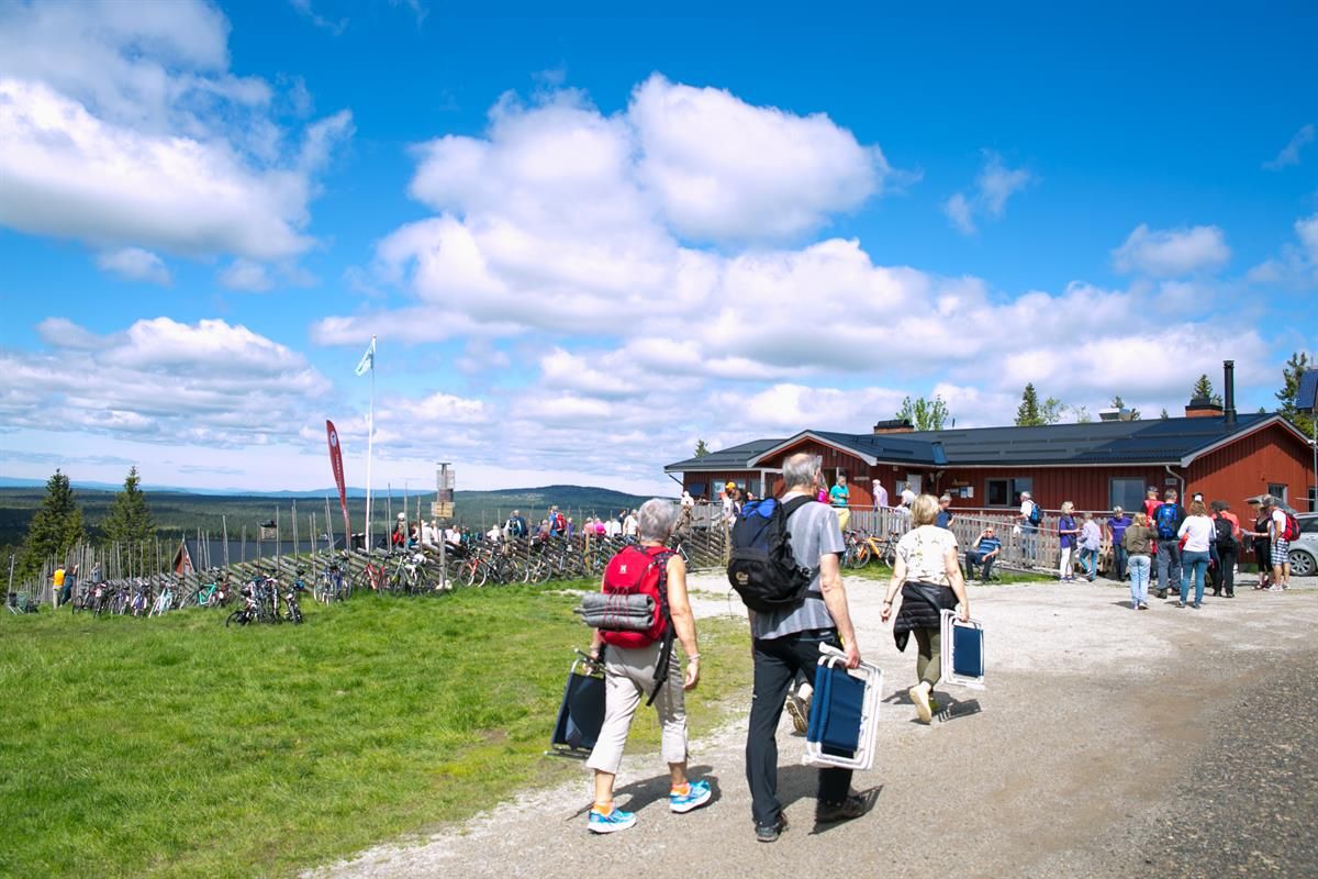 Folk beveger seg rundt uteområdet ved serveringshytta Målia, omgitt av gress, sykler og klar blå himmel.
