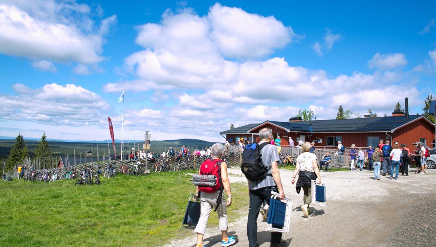 Folk beveger seg rundt uteområdet ved serveringshytta Målia, omgitt av gress, sykler og klar blå himmel.