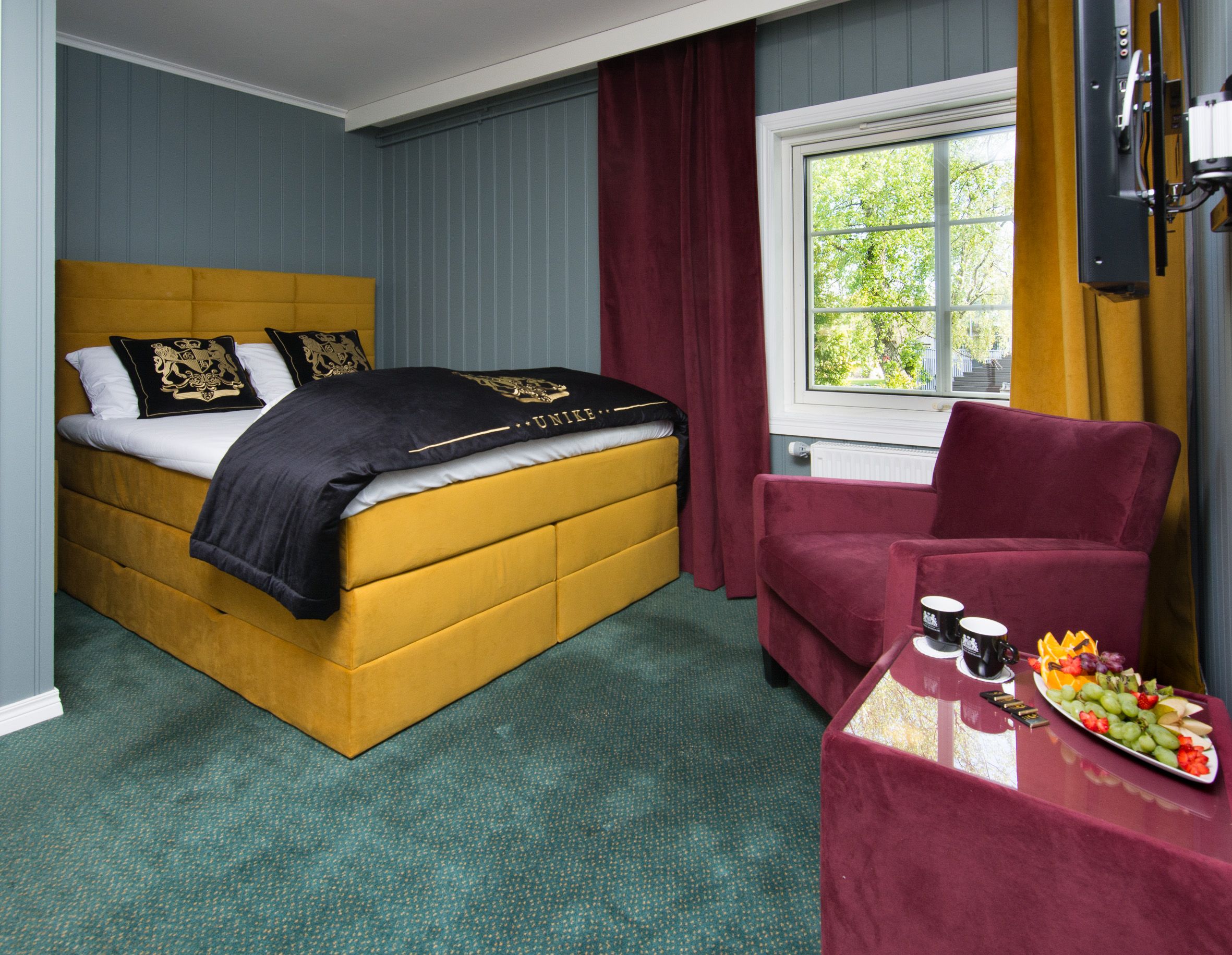 dobbeltrom med gul seng, grønt teppe og vinrød fåtølj.
