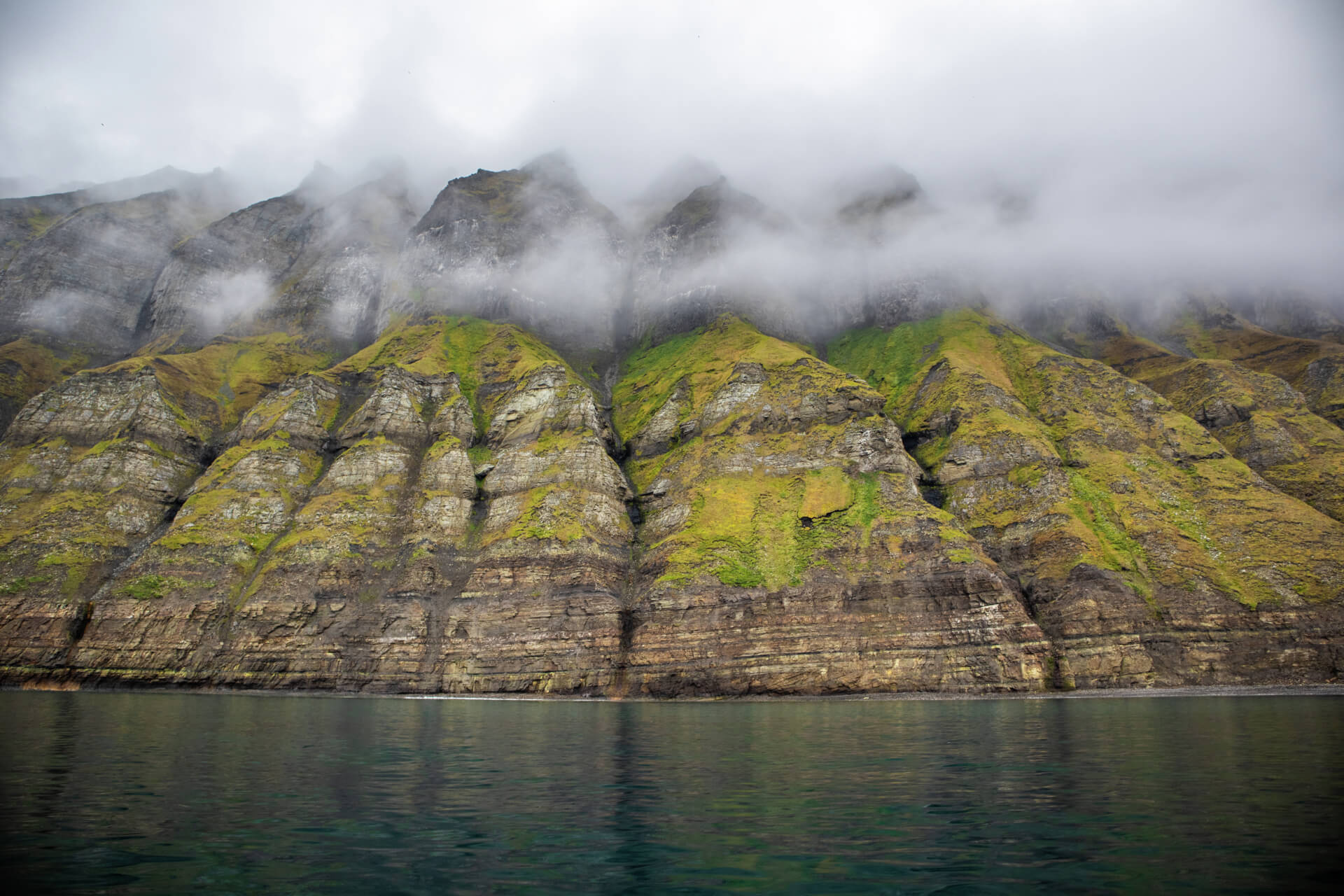 Massive green mountains rising up next to a fjord with mist in between the mountain's peaks