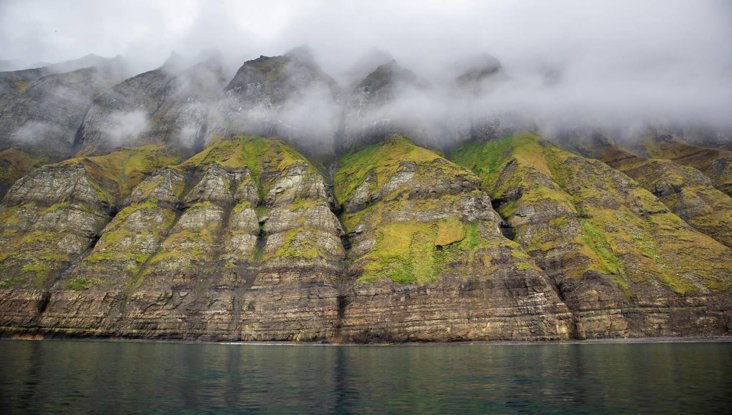Massive green mountains rising up next to a fjord with mist in between the mountain's peaks