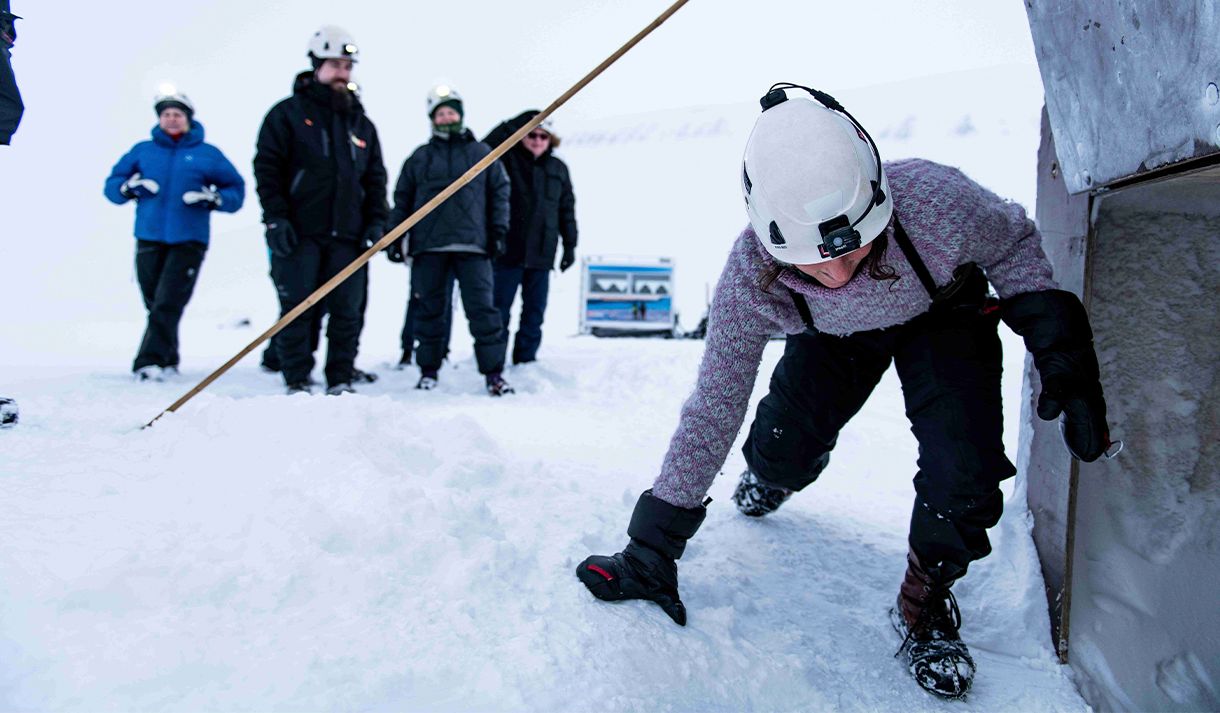 A guide with a headlamp and helmet climbing down into the entrance to an ice cave in the foreground, with guests waiting their turn in the background