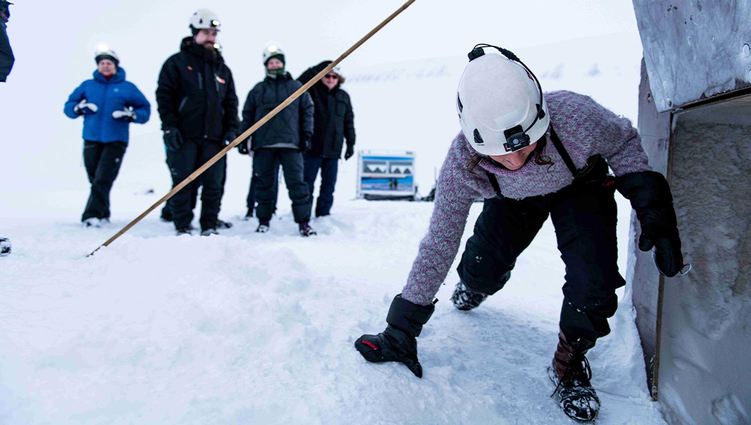 A guide with a headlamp and helmet climbing down into the entrance to an ice cave in the foreground, with guests waiting their turn in the background