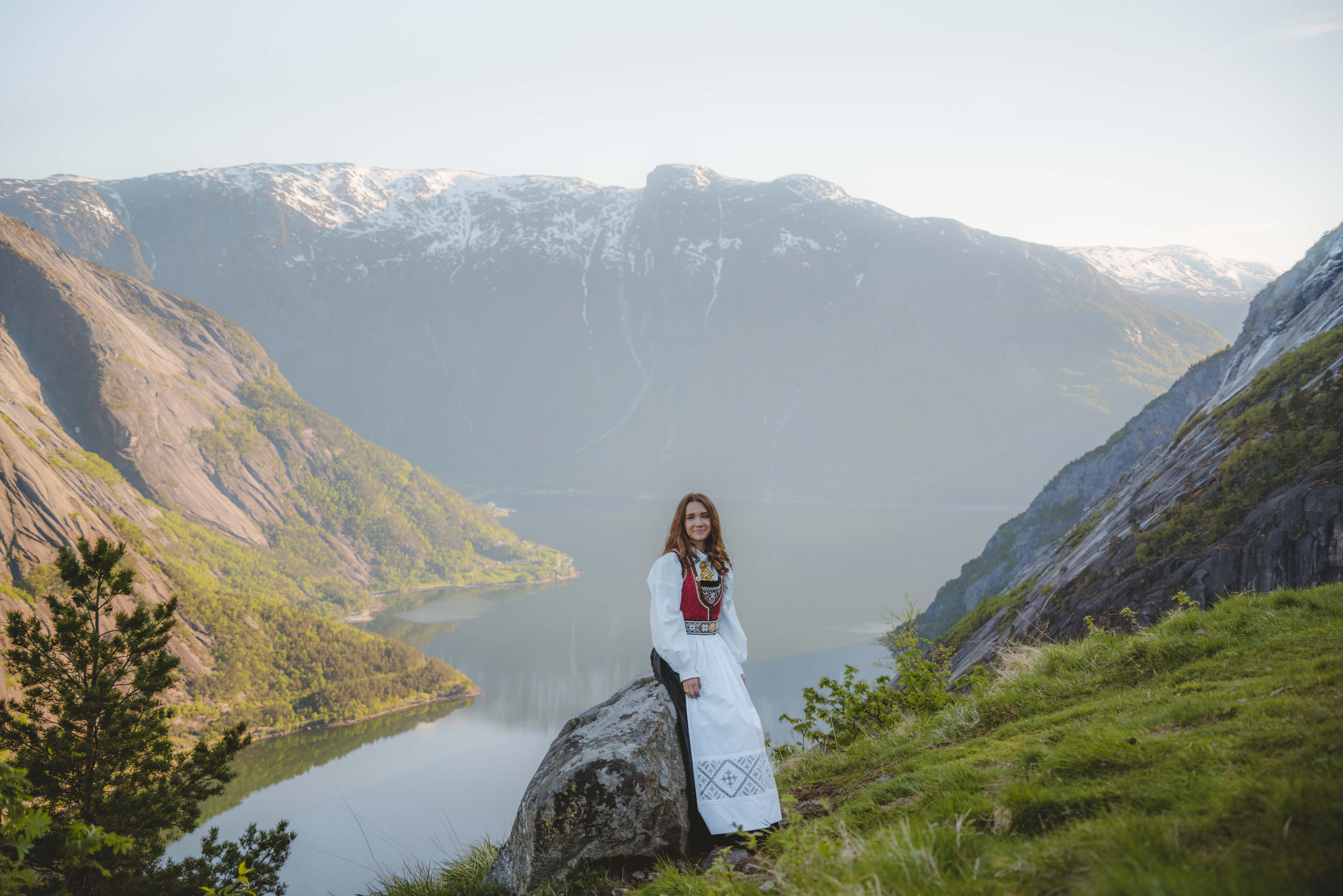 En jente iført bunad, sittende på en stein med fjorden og fjellene i bakgrunnen.