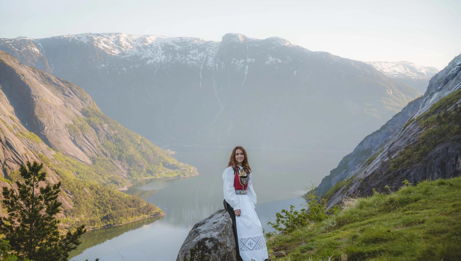 En jente iført bunad, sittende på en stein med fjorden og fjellene i bakgrunnen.