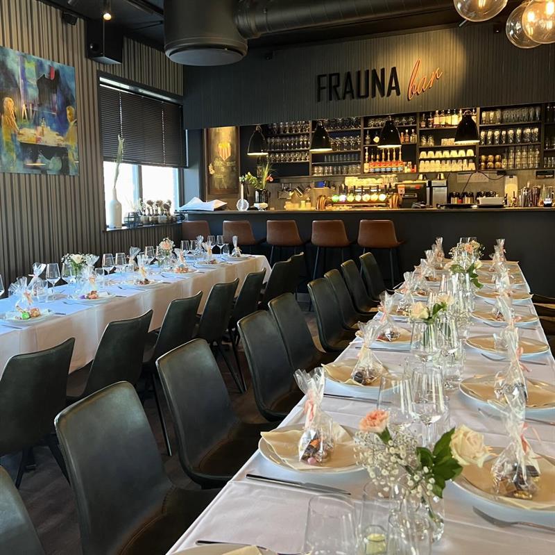 Dekket langbord inne i restauranten med glass, servietter og blomsterpynt. Vinhyller og mørke vegger skaper en elegant og intim atmosfære.