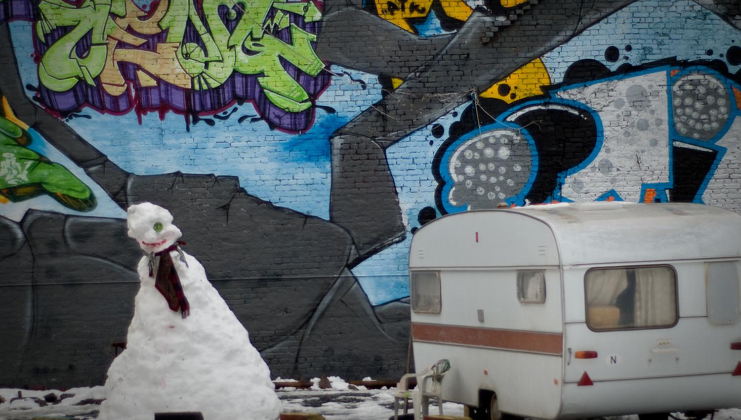 En snømann og en campingvogn foran en vegg med grafitti.