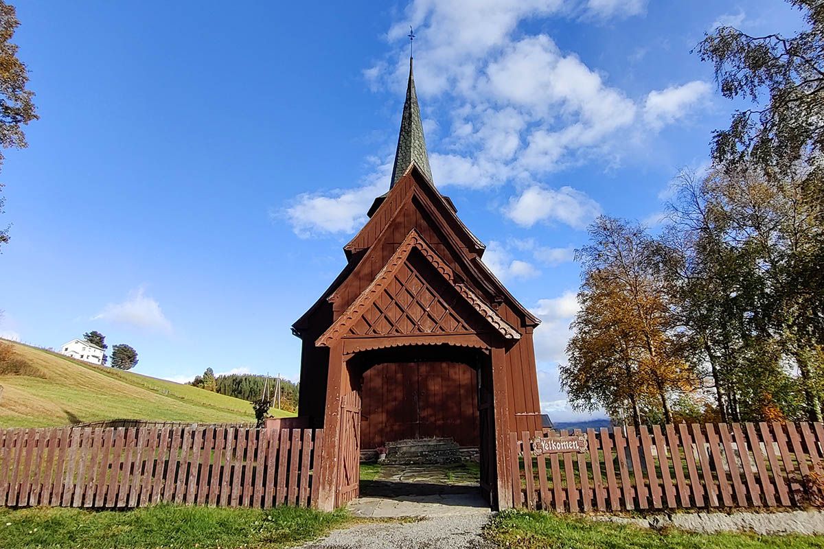 A stave church beyond  a wooden fence with a portal with ornaments. Blue skies.