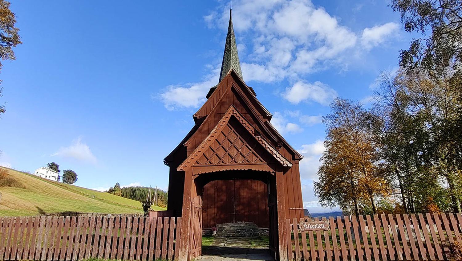 A stave church beyond a wooden fence with a portal with ornaments. Blue skies.