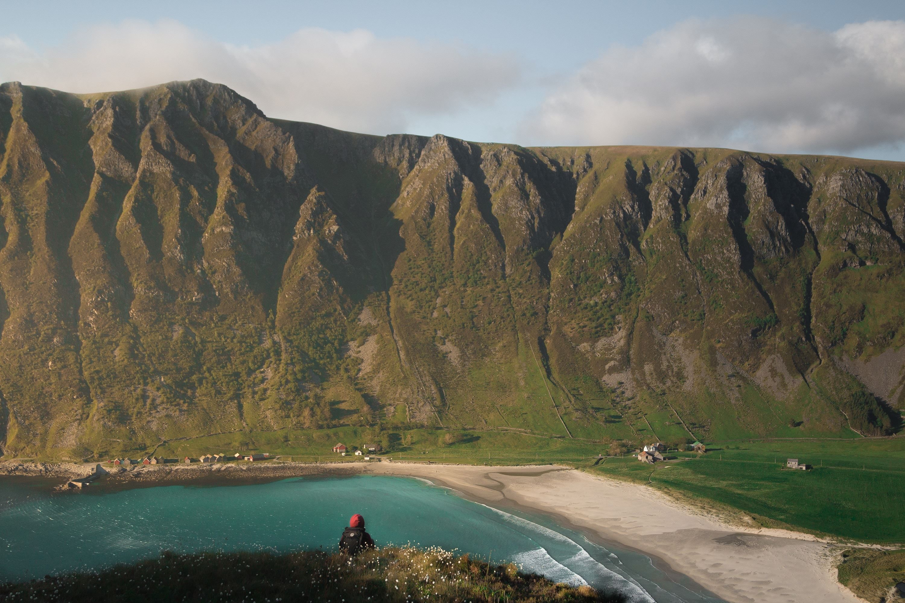A lone hiker sits on a grassy cliff overlooking a turquoise bay, sandy beach, and steep green mountains under clouds..