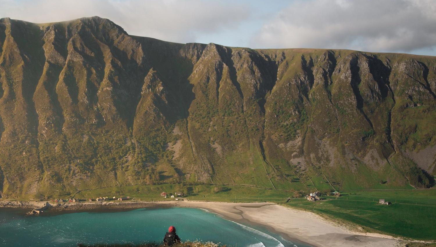 A lone hiker sits on a grassy cliff overlooking a turquoise bay, sandy beach, and steep green mountains under clouds..