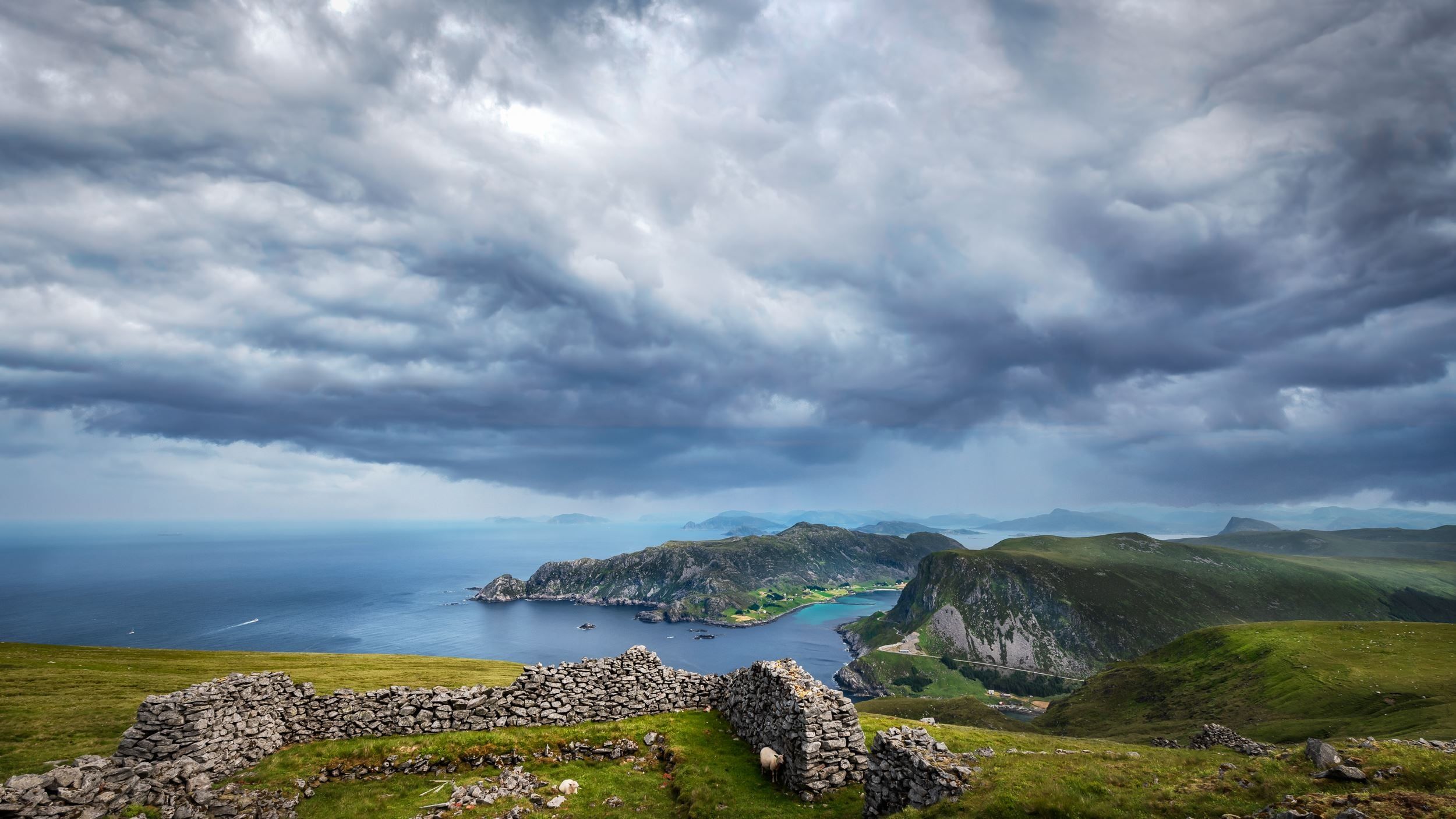 Utsyn frå grøn høgde med steinmur mot ei fjordliknande vik, bratte fjell og mørke stormskyer over havet..