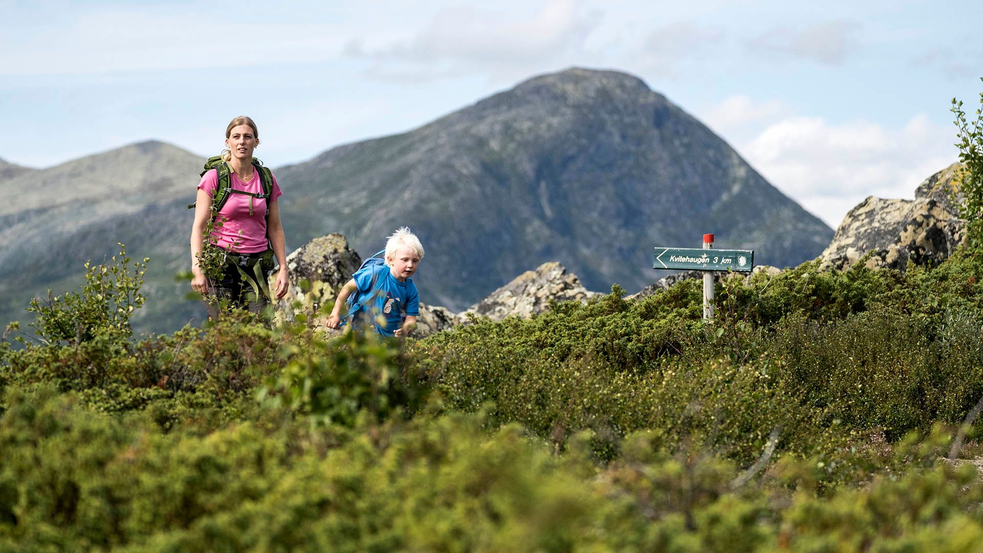 Ung kvinne med gutt på tur langs sti med einebærbusker i forgrunnen. I bakgrunnen ses fjell.