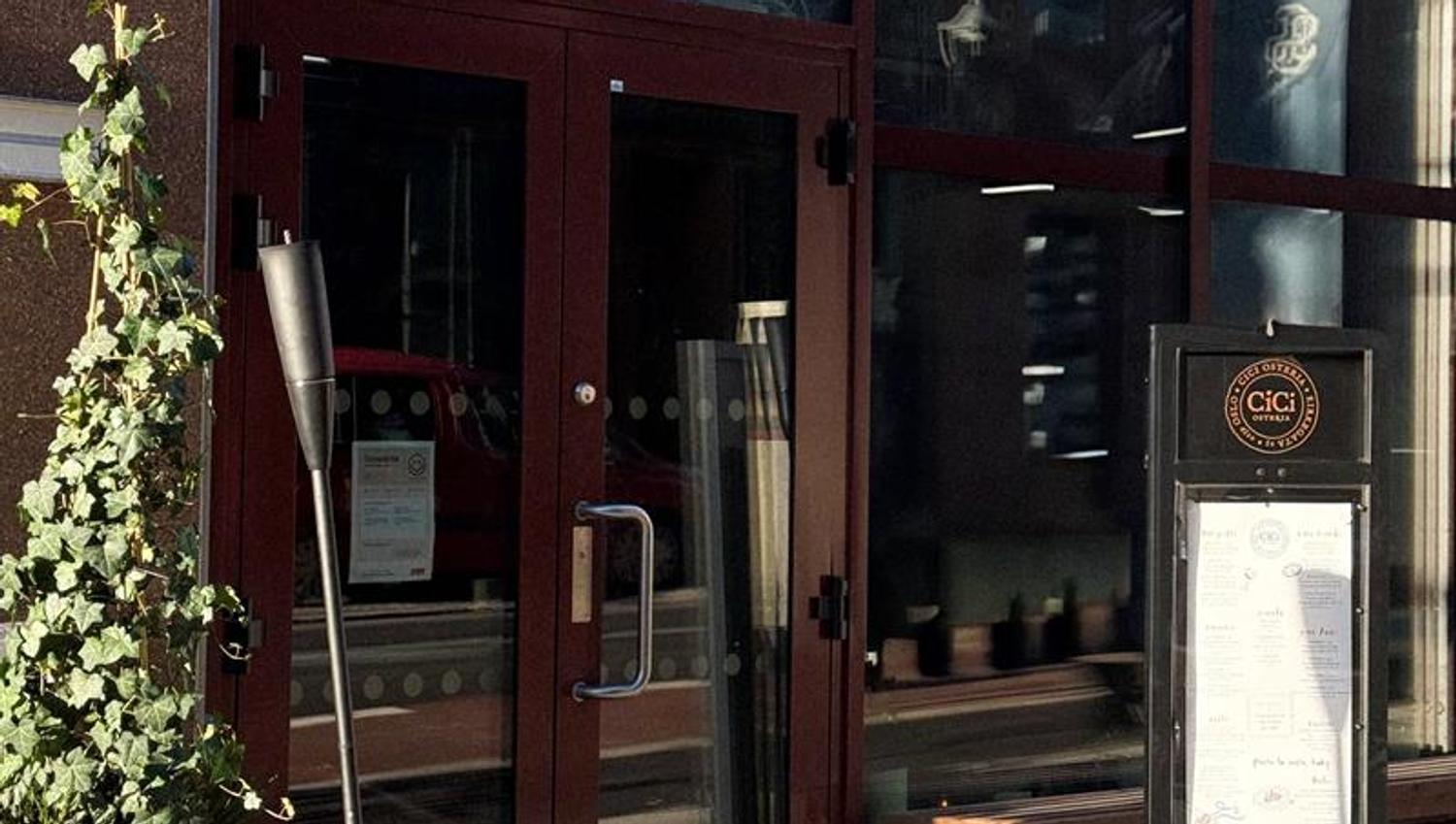 The entrance to a restaurant with large glass windows, a menu display by the door, and a lantern and plant placed on the pavement.