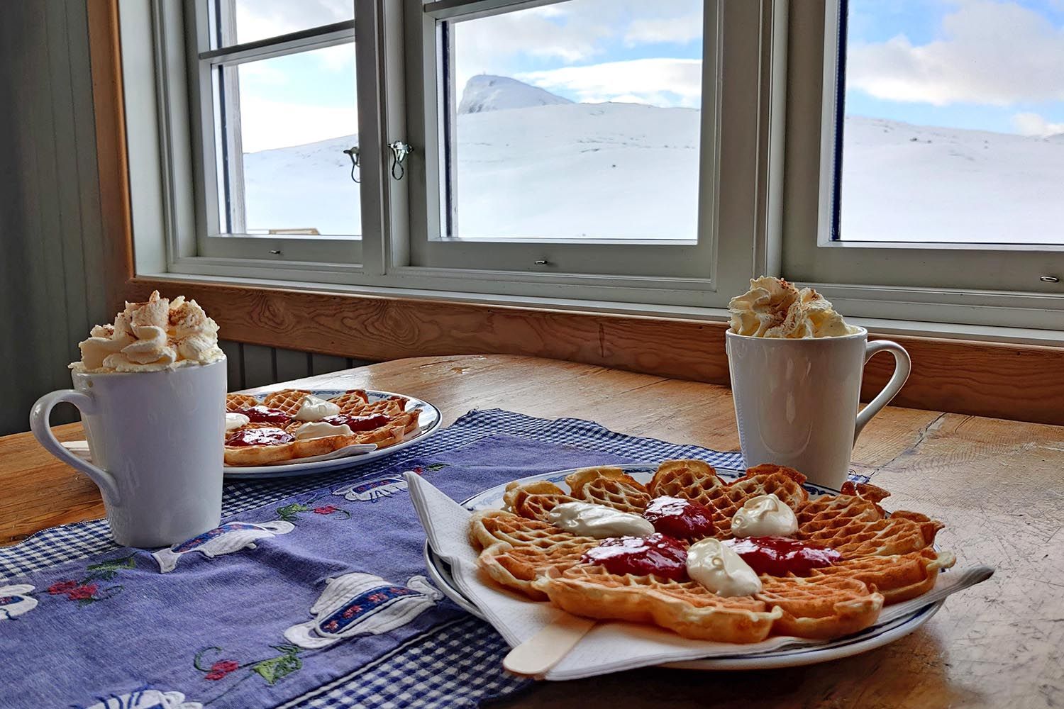 Vaffler med rømme og syltetøy, kakao på krus med krem og utsikt mot vinterlige fjell.