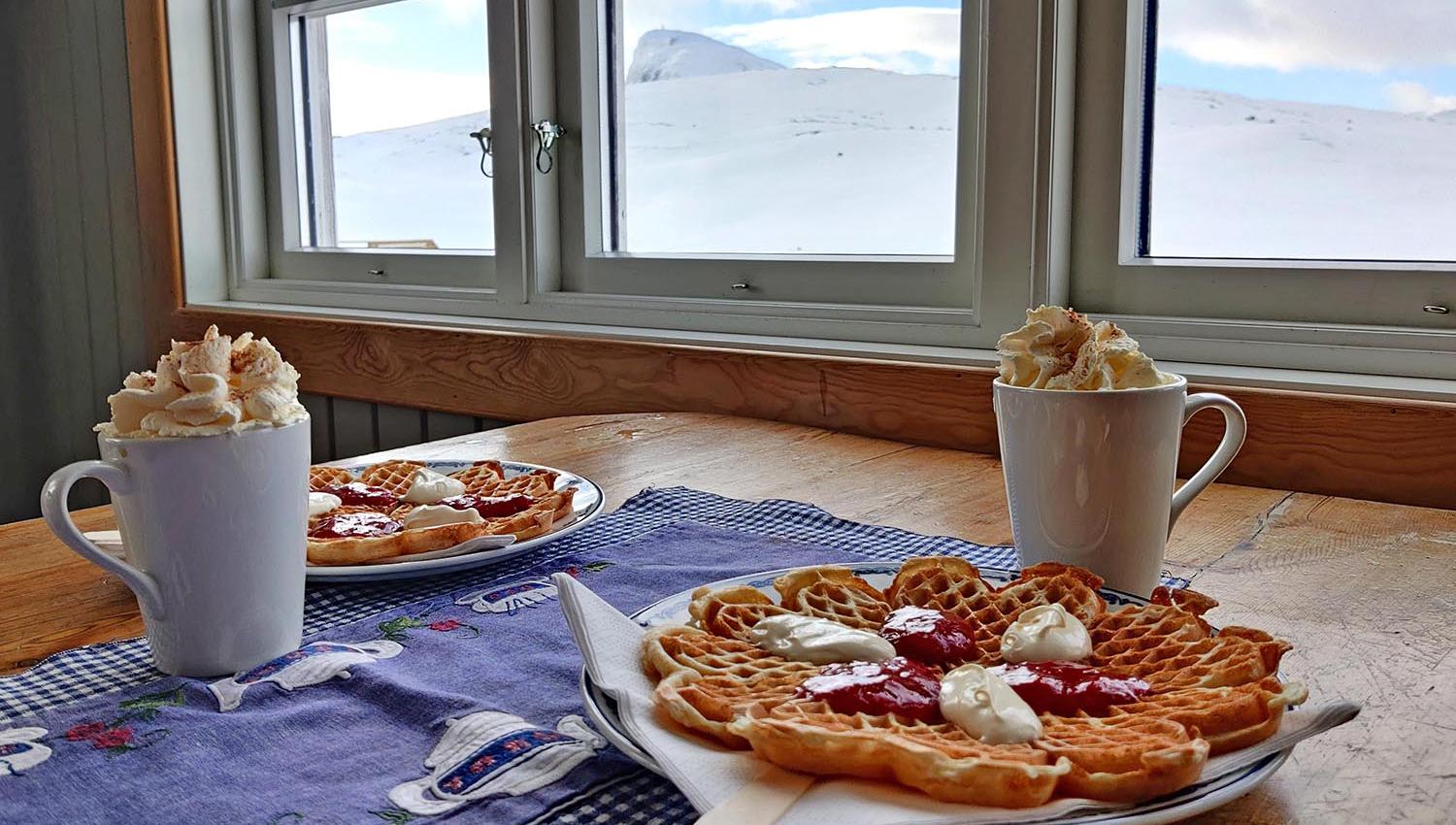 Vaffler med rømme og syltetøy, kakao på krus med krem og utsikt mot vinterlige fjell.