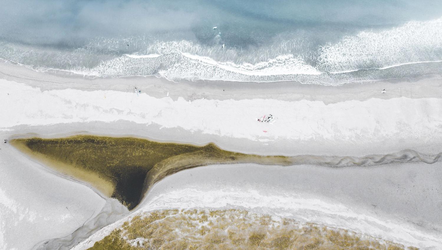 Luftfoto av bølgjer som slår mot ein lys sandstrand, med eit mørkare elvelaup og grasområde i framkant..