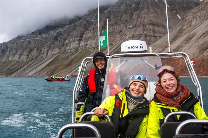 Tre personer smiler på RIB-båt med fjell i bakgrunnen.